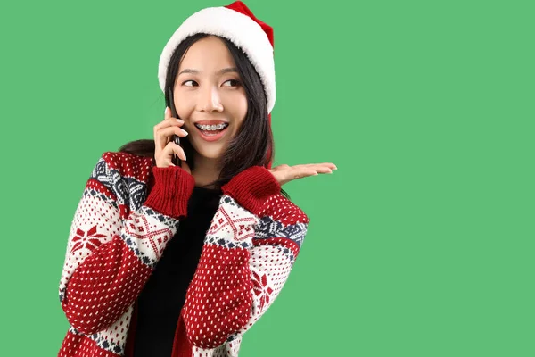 サンタハットの若い美しいアジアの女性 緑の背景で携帯電話で話す — ストック写真