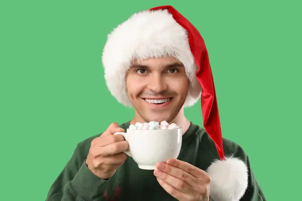 快乐的年轻人戴着圣诞礼帽 拿着一杯绿底棉花糖的美味可可 — 图库照片