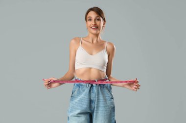 Gri arka planda belini ölçen bol kotlu genç bir kadın. Kilo verme konsepti