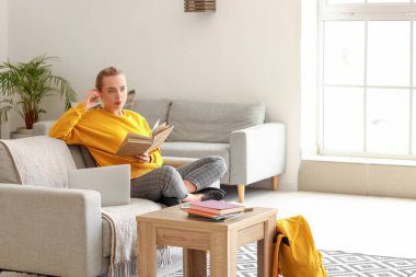 Bayan öğrenci evdeki kanepede kitap okuyor.