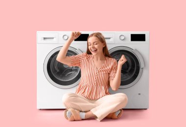 Mutlu genç kadın pembe arka planda çamaşır makinelerinin yanında oturuyor.