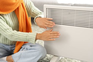 Genç bir kadın, kaloriferin yanında ellerini ısıtıyor.