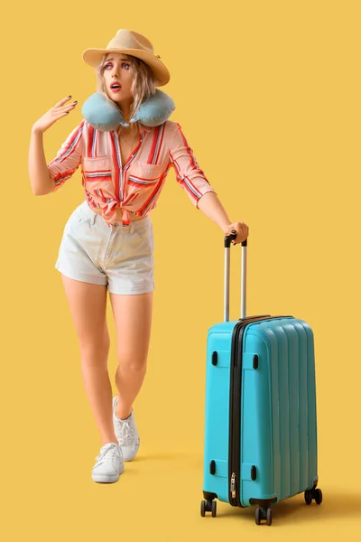 Young woman with neck pillow and suitcase on yellow background