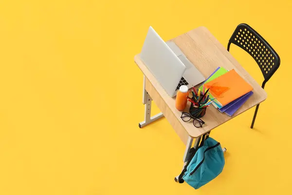 Modern school desk with backpack, laptop and stationery on yellow background