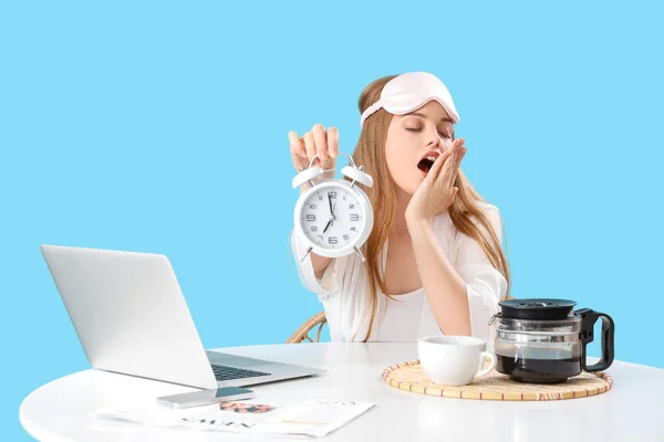 Sleepy young woman with alarm clock and laptop at table on blue background