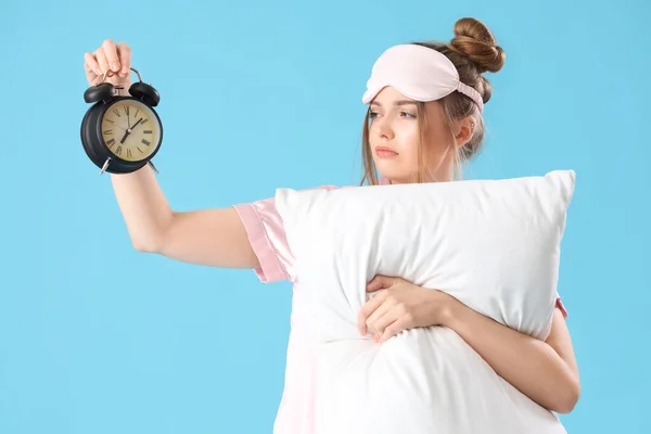 Sleepy young woman with alarm clock and pillow on blue background