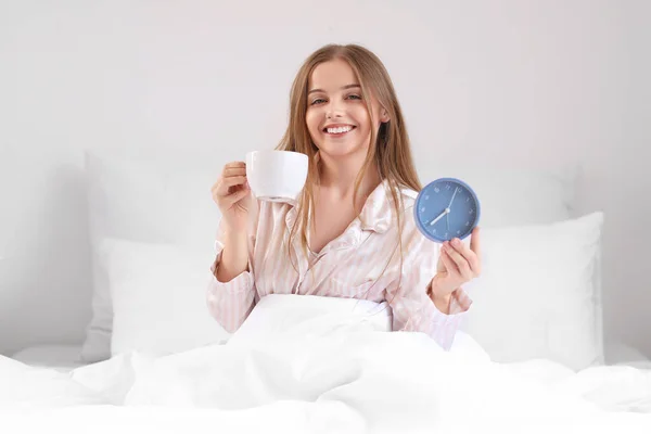 自宅でベッドでコーヒーとアラームクロックのカップを持つ若い女性 — ストック写真