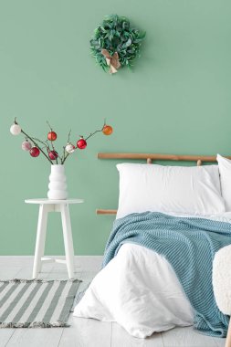 Yeşil duvarın yanındaki dallarda Noel topları olan rahat bir yatak ve vazo.