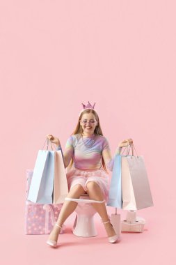 Alışveriş torbalı genç bir kadın pembe arka planda klozetin üzerinde oturuyor.