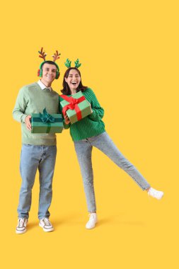 Mutlu genç çift Noel geyiği boynuzlarıyla hediye kutularıyla sarı arka planda