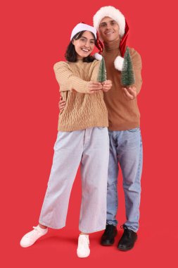 Noel Baba şapkalı, kırmızı arka planda köknar ağaçları olan mutlu bir çift.