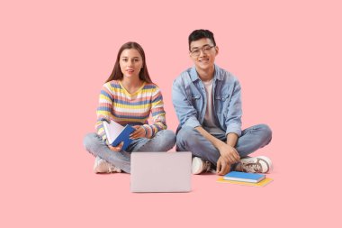 Kitapları pembe arka planda oturan ve dizüstü bilgisayar kullanan mutlu öğrenciler.