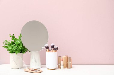 Odadaki pembe duvarın yanındaki tuvalet masasında farklı makyaj malzemeleri, ayna ve bitkiler var.