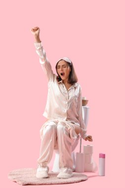 Pembe arka planda pijamalı, klozetin üzerinde oturan uykulu genç kadın.