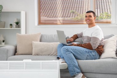 Evde radyatörün yanında laptoplu genç adam ısınıyor.