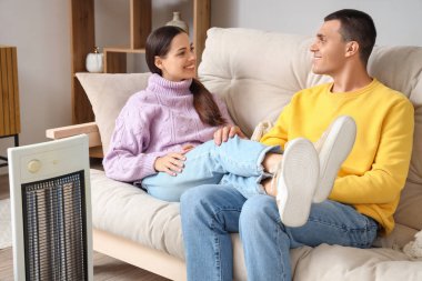 Evde elektrikli ısıtıcının yanında ısınan genç bir çift.