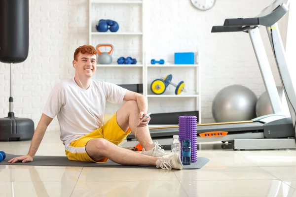 Sporty redhead man with earphones and mobile phone listening to music in gym