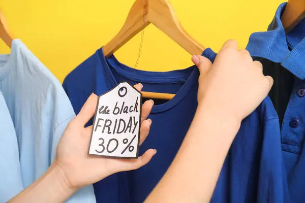 Female hands with sale tag and t-shirts against yellow background, closeup. Black Friday