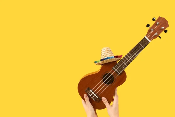 Female hands holding ukulele with sombrero on yellow background