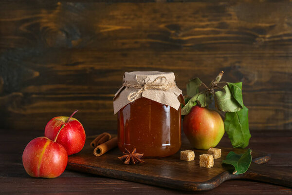 Cutting board with jar of sweet apple jam and ingredients on wooden background