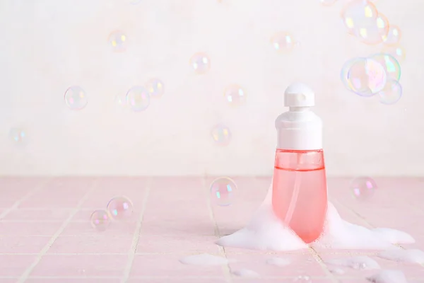 Bottle of cosmetic product with foam and soap bubbles on color tile table against light background