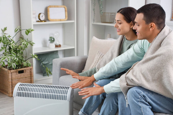Young couple with plaid warming hands near radiator at home