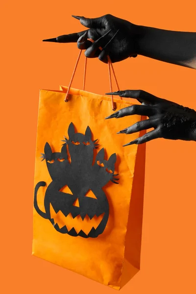 Black hands of witch with Halloween bag on orange background