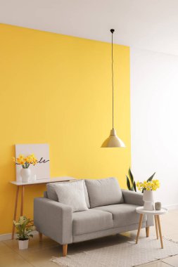 Oturma odasının içinde gri kanepe ve kahve masasında çiçek açan narsis çiçekleri.