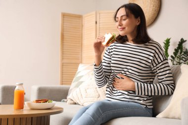 Oturma odasındaki kanepede dinlenirken lezzetli sandviçler yiyen güzel genç bir kadın.