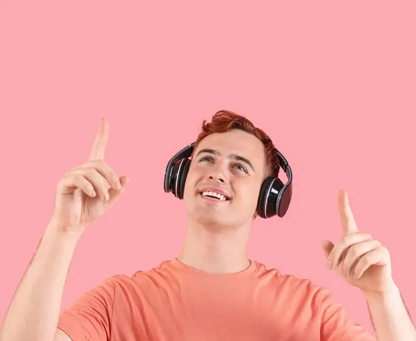 ピンクの背景に何かを指すヘッドフォンの幸せな若い赤毛の男 — ストック写真