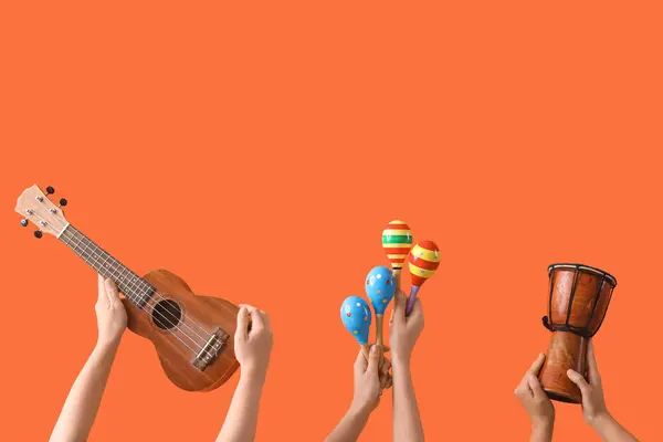 Female hands holding ukulele, drum and maracas on orange background