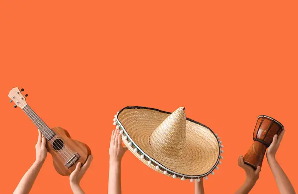 Female hands holding sombrero, ukulele and drum on orange background