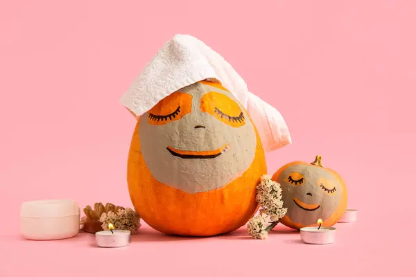 Pumpkins with clay mask, cosmetic products and burning candles on pink background