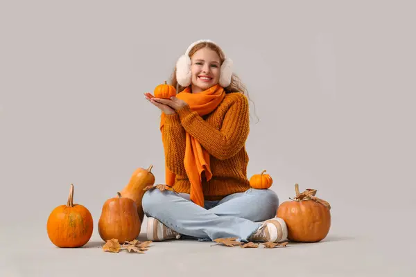 Young woman with pumpkins and autumn leaves sitting on light background