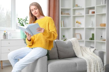 Evdeki kanepede kitap okuyan genç bir kadın.