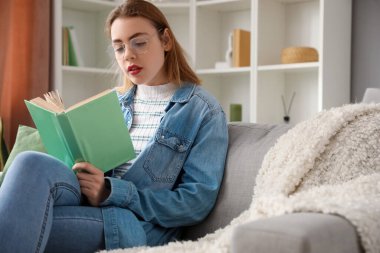 Evdeki kanepede kitap okuyan genç bir kadın.