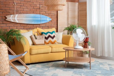 Sarı kanepesi, sörf tahtası ve kahve masasında elektrikli vantilatörü olan şık bir oturma odası.