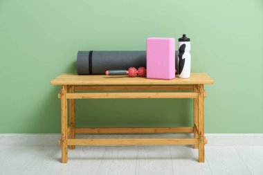 Yoga minderli ahşap masa, blok, masaj silindiri ve yeşil duvarın yanında bir şişe su.