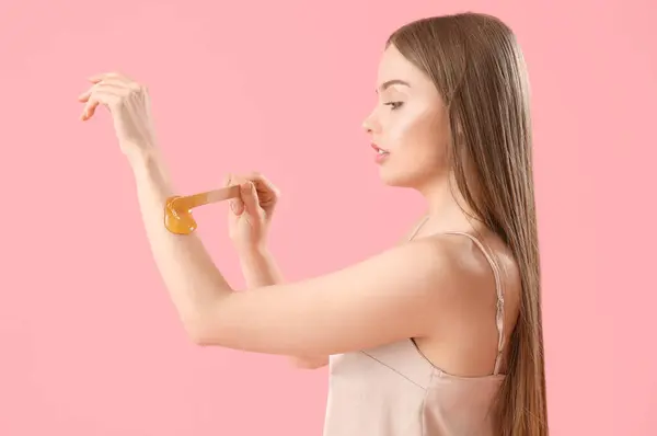 Young woman applying sugaring paste onto her arm against pink background