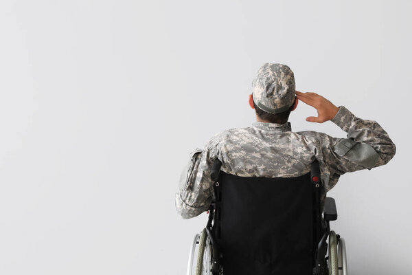 Молодой солдат в инвалидной коляске салютует на светлом фоне, вид сзади