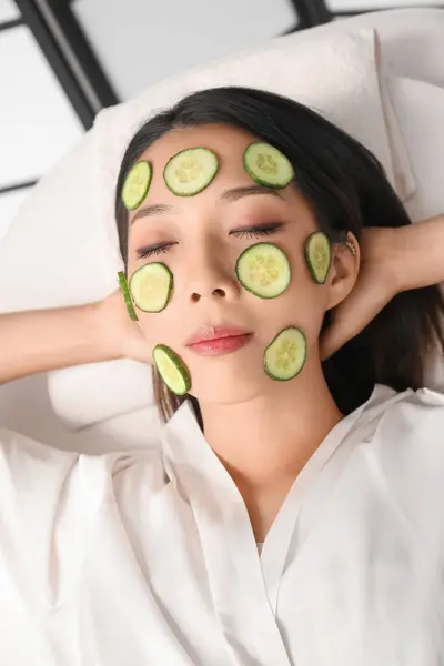 Young Asian woman with cucumber slices relaxing in spa salon, closeup