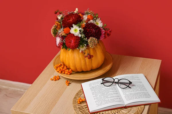 Beautiful autumn bouquet in pumpkin and book with eyeglasses on wooden table against red background