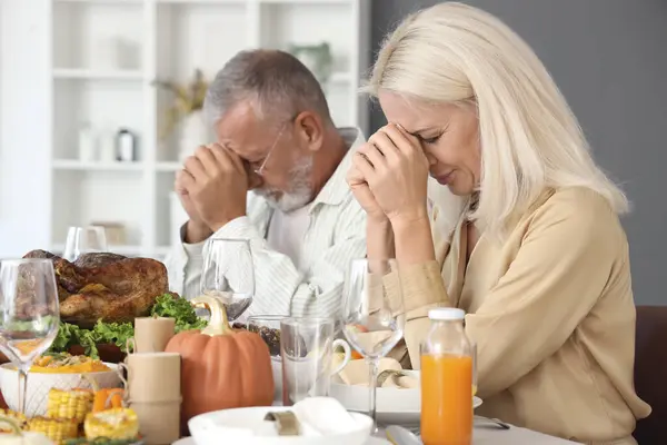 Mature couple praying before dinner at festive table on Thanksgiving Day