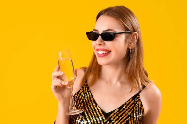 Güneş gözlüklü, sarı arka planda bir kadeh şampanyası olan güzel bir kadın.