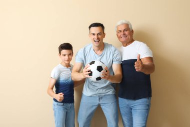 Babası ve dedesiyle birlikte bej arka planda futbol topu tutan küçük bir çocuk.