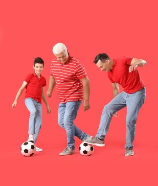 Babası ve dedesiyle kırmızı arka planda futbol oynayan küçük bir çocuk.