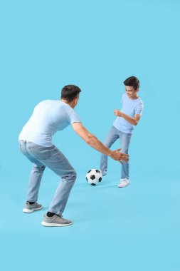 Küçük çocuk ve babası mavi arka planda futbol oynuyorlar.