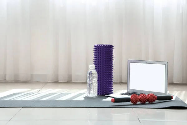 Laptop with bottle of water and rollers on fitness mat in gym