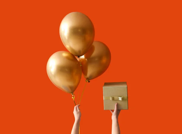 Female hands with golden balloons and gift for birthday celebration on color background