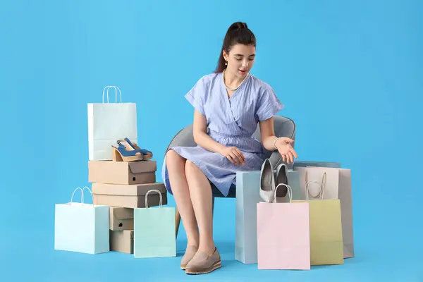 Young woman with shoe boxes and shopping bags sitting in armchair on blue background